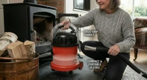 A photorealistic illustration showing a person lifting the compact red ash vacuum by its integrated black carry handle, set on a slate hearth.