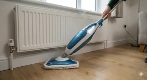 A steam mop with a swivel head cleaning around the base of a radiator and kitchen units on a laminate floor.