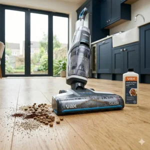 A woman using a modern floor cleaner to clean near kitchen cabinets with a dog resting nearby.