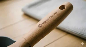 A hand brush sweeping small leaves and dirt particles into a grey dustpan on a light-coloured wooden kitchen floor.